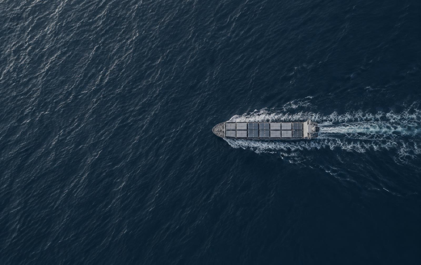 Navio cargueiro visto de cima atravessando o oceano, deixando um rastro branco